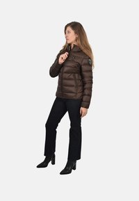 Hnedá zateplená puffer bunda s horizontálnym prešívaním, so zapínacími vreckami a logom na ľavom pleci; nosená s tmavými džínsami a ihličkovými topánkami.