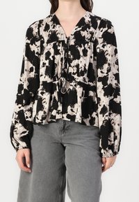 Schwarz-beige gemusterte Bluse mit langen Ärmeln, gerafftem Bund und V-Ausschnitt. Das Material wirkt weich mit einem fließenden Design.