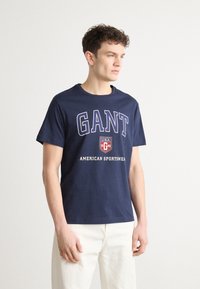 Tricou din bumbac bleu marin, cu text alb îndrăzneț „GANT” și „AMERICAN SPORTSWEAR”, guler rotund și mâneci scurte. Design simplu.