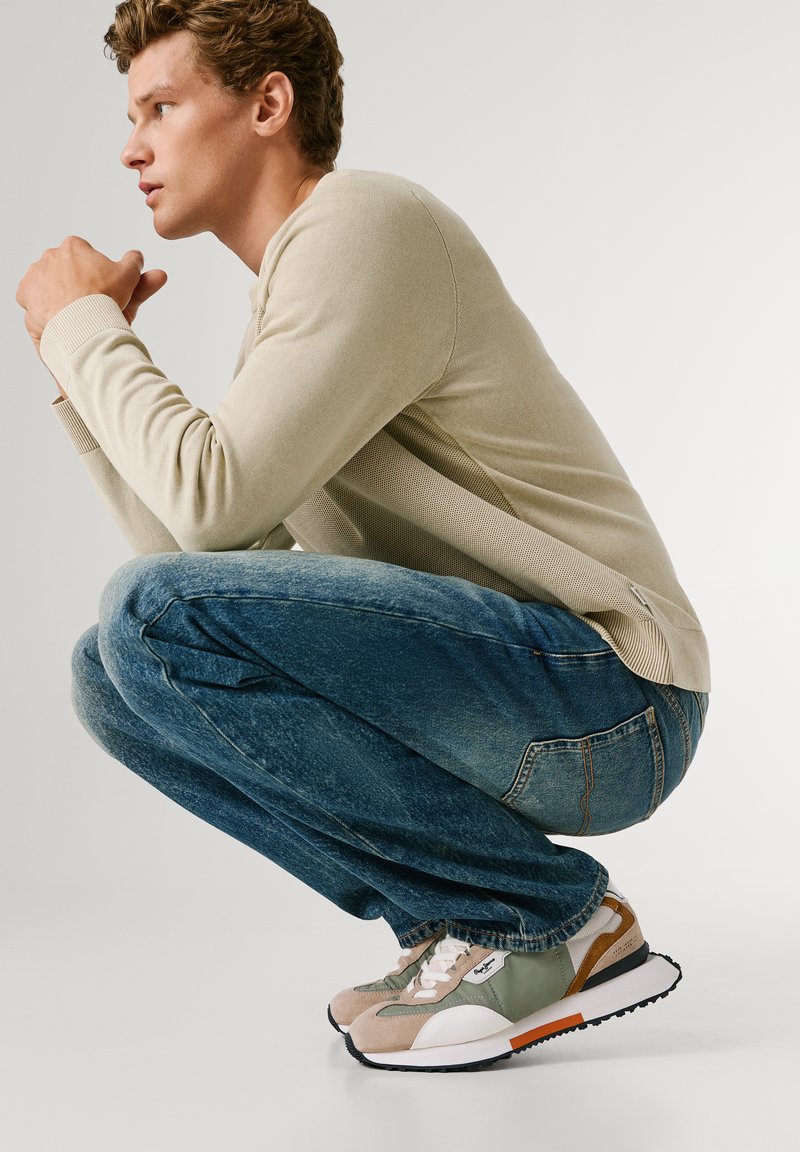 Młody mężczyzna kucający z splecionymi dłońmi, ubrany w beżowy sweter, niebieskie dżinsy oraz zielone, beżowe i białe trampki na jasnym tle.
