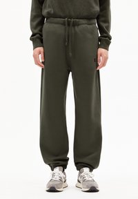 Olijfgroene joggers met een trekkoord in de taille, elastische boorden, zachte stof en subtiele logo details. Gecombineerd met grijze sneakers.