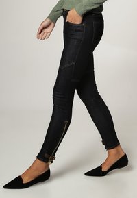 Úzké tmavé džínové kalhoty se zlatým stehem, které mají zipy na kotnících a přezkové detaily. Nosené s černými špičatými balerínami.