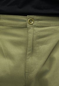 Artimųjų planas alyvuogių žalių kelnių su sagos užsegimu ir matomomis siūlėmis, dėvėtų su juoda palaidine.