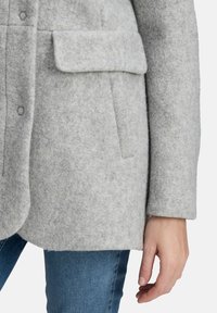 Cappotto grigio in misto lana con tasca a pattina, chiusura con bottoni e finitura testurizzata; abbinato a jeans blu.