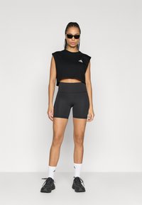 Μαύρο, κοντό αθλητικό τοπ με κοντά μανίκια, σετ με ψηλόμεσο μαύρο σορτς. Αξεσουάρ περιλαμβάνουν γυαλιά ηλίου και άσπρες κάλτσες.