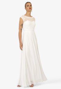 Witte lange jurk met een kanten lijfje en kapmouwen, met een geraffineerde taille en een vloeiende rok. Model draagt subtiele accessoires.