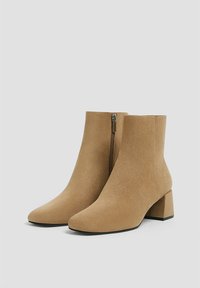 Bottines en daim beige avec un bout fermé, un talon bloc et une fermeture éclair sur le côté. La surface a une texture lisse, avec des détails de couture minimaux.