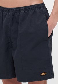 Mørkeblå shorts laget av lett stoff, med elastisk midje, sidelommer og en brodert logo i oransje på kanten.