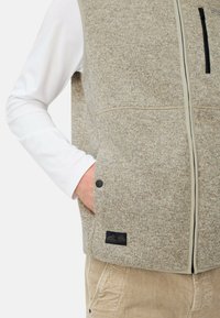 Beige strukturiertes Weste mit Reißverschluss vorne, Seitentaschen und einem schwarzen Akzent-Tag. Über einem weißen Langarmshirt getragen. Cord-Hosen sichtbar.