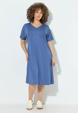 Kvinde med krøllet hår iført en knælange blå kjole med korte ærmer og hvide sneakers, stående mod en ensfarvet hvid baggrund.