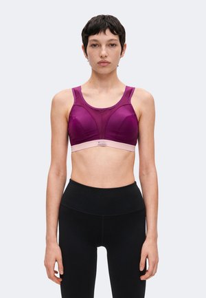 Ung person med kort mørkt hår iført en lilla sports-bh og sorte leggings med høj talje, stående mod en ensfarvet baggrund.