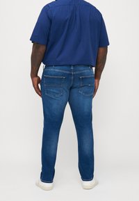 Man som bär en blå kortärmad skjorta, blå jeans med bakfickor och vita sneakers, stående vänd bort från kameran mot en enkel bakgrund.