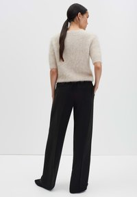 Femme avec une longue queue de cheval noire portant un pull en beige moelleux à manches courtes et un pantalon noir à jambes larges, se tenant de dos.