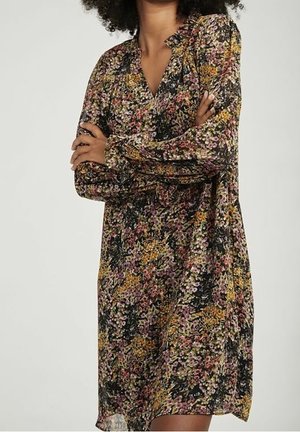 Osoba w długiej sukience w kwiaty w różowe, żółte i zielone wzory, z założonymi skrzyżowanymi ramionami, na jednolitym jasnym tle.