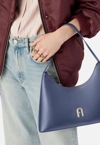 Niebieska skórzana torebka o zaokrąglonym kształcie, z logo z złotym okuciem. Nosi się ją swobodnie z bordową kurtką bombową i jasnymi dżinsami.