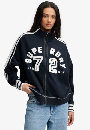 Kvinde med langt mørkt hår har en marineblå lynlåstrøje med hvide striber og store hvide "Superdry 72" bogstaver på, kombineret med lyseblå jeans.