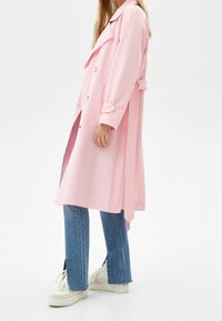 Long pink trench coat with a loose fit, large collar, and button closure. Paired with light blue jeans featuring slits and white sneakers.