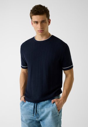 Mann trägt ein dunkelblaues, kurzes Strickshirt mit weißen Absetzungen, hellblaue Jeans und eine silberne Gliederkette, die Hände in den Taschen.