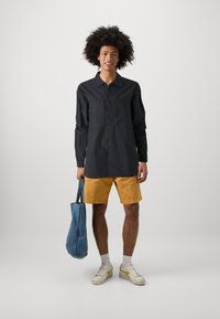 Black button-up shirt with long sleeves, paired with yellow shorts. Holding a blue tote bag. Wearing white sneakers with light-colored socks.