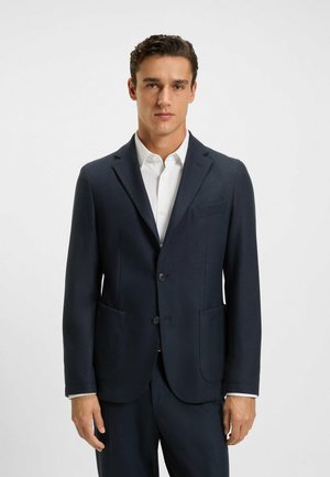 Uomo che indossa una giacca da abito sartoriale blu scuro su una camicia bianca con bottoni, in piedi davanti a uno sfondo chiaro e uniforme.