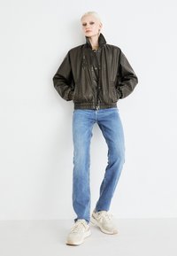Olive green leather bomber jacket with a high collar, zippered chest pocket, and elasticized cuffs, paired with light blue jeans and beige sneakers.