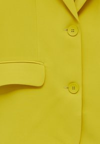 Gros plan sur un blazer jaune montrant deux boutons ronds et une poche à rabat sur un tissu lisse.