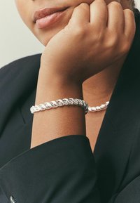 Silver chain bracelet with a twisted design, worn on a wrist, showcasing a shiny, reflective surface against a black clothing background.