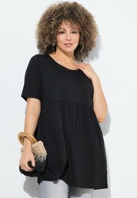 Černý top s krátkým rukávem a volnějším střihem, s nařaseným pasem. Modelka drží malou tkanou tašku s dřevěnými držadly.