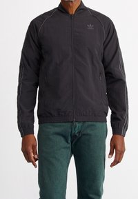 Black zip-up jacket with thin grey stripes on sleeves. Features a standing collar and two side pockets. Material appears lightweight and textured.