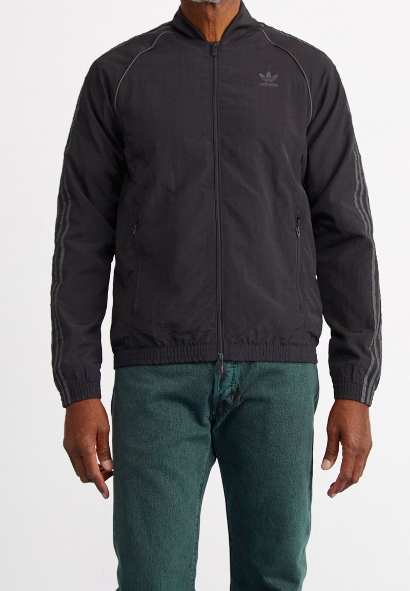 Black zip-up jacket with thin grey stripes on sleeves. Features a standing collar and two side pockets. Material appears lightweight and textured.