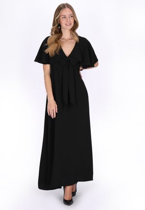 Vrouw met los golvend haar, gekleed in een lange zwarte jurk met zwierige mouwen en een strik aan de voorkant, staand en lichtjes zijwaarts kijkend.