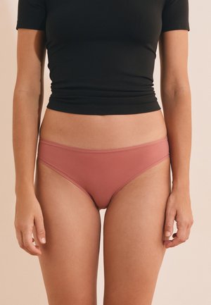 Frau trägt enganliegendes schwarzes Kurzarmtop und rosafarbene nahtlose Bikini-Unterwäsche, steht vor einem beigen Hintergrund.