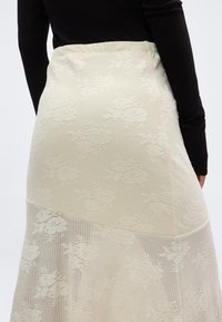 Γυναίκα που φοράει midi φούστα από κρεμ δαντέλα με λουλούδια και μαύρη μπλούζα μακρυμάνικη, εμφανιζόμενη από τη μέση πίσω μέχρι το μέσο του μηρού.