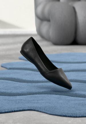 Chaussure plate en cuir noir à bout pointu flottant au-dessus d'un tapis bleu texturé avec un canapé gris flou en arrière-plan.
