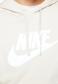 Béžová mikina s kapucí z měkké látky, zdobená výrazným bílým logem Nike a proužkem swoosh. Součástí jsou také šňůrky na kapuci.