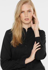Black button-up shirt with a collar, featuring long sleeves and a smooth texture. Notable details include buttons and a label at the hem.