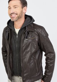 Ruskea nahkatakki, jossa on tuftattuja olkasuojusta, vetoketjullisia taskuja ja harmaa huppari alla. Sileä pinta, moderni muotoilu.