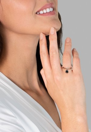 Anello d'argento con una piccola pietra nera, indossato su una mano che tocca il mento. Texture liscia, design minimalista, su uno sfondo neutro.