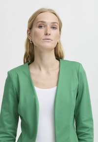 Zöld blézer sima anyagból, testhez álló fazonnal és V-nyakkal, fehér felsőre rétegezve. Egyszerű és letisztult sziluett.