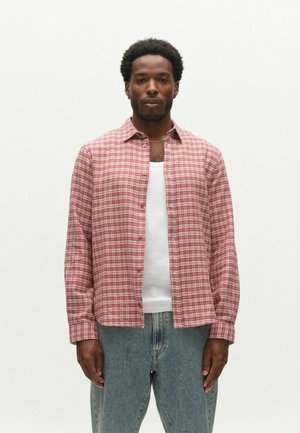Man wearing a red and white checkered button-up shirt over a white tank top and blue jeans, standing against a plain light background.