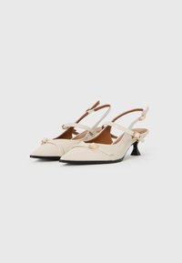 Kengät koralliväristä patenttilahkeista slingback-mallia, joissa on teräväkärkiset varpaat, kultaiset solkiyksityiskohdat ja mustat matalat liehuvat korot valkoista taustaa vasten.