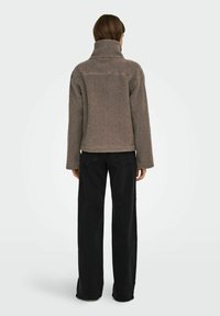Fleece bunda v taupe barvě, s vysokým límcem, volným střihem a strukturovaným povrchem, spárovaná s černými kalhotami širokého střihu.