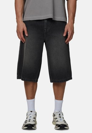 Osoba ubrana w wyblakłe czarne długie szorty, białe krótkie skarpetki oraz biało-czarne buty sportowe, stojąca z rękami swobodnie opartymi po bokach.