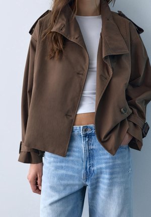 Personne portant une veste marron oversize, un top blanc court et un jean taille haute bleu clair, debout devant un fond uni.