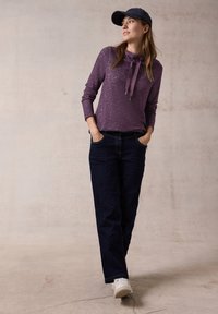 Lila langärmeliger Kapuzenpullover mit Schnürung, kombiniert mit dunkelblauen Gerade-Passform-Jeans und weißen Sneakern. Lässiges Outfit vor einem neutralen Hintergrund.