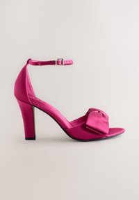 Roze satijnen sandaal met hoge hak, open teen, enkelbandje en grote strik op het voorblad. Gladde textuur en glanzende afwerking.