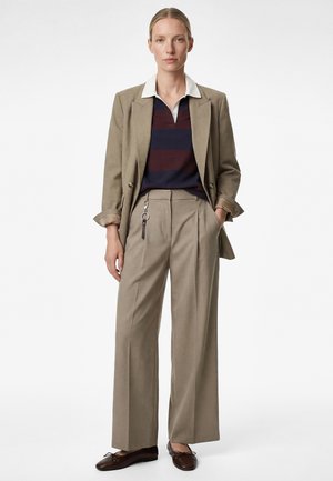 Kobieta ubrana w beżową, dopasowaną marynarkę i szerokie spodnie, w granatowo-bordową prążkowaną koszulkę polo i brązowe baleriny, stojąca z rękami w kieszeniach.