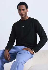 Juodi ilgomis rankovėmis marškinėliai su balta „BOSS“ logotipu krūtinės srityje, derinami su šviesiai mėlynomis kelnėmis. Lygus audinys, laisvas siluetas ir paprastas dizainas.