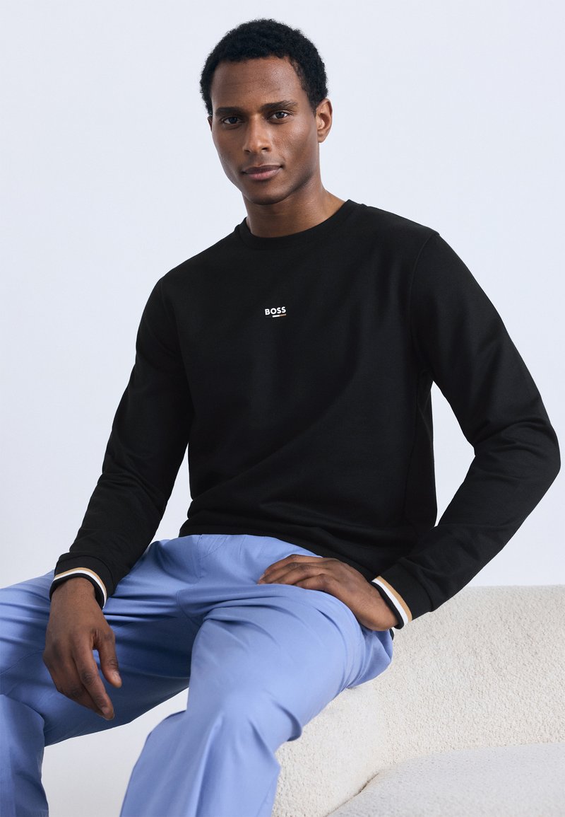 Juodi ilgomis rankovėmis marškinėliai su balta „BOSS“ logotipu krūtinės srityje, derinami su šviesiai mėlynomis kelnėmis. Lygus audinys, laisvas siluetas ir paprastas dizainas.