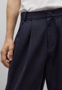 Pantalones plisados de color azul marino con tela texturizada y diseño de frente plano. Cuentan con bolsillos laterales y cintura con trabillas para cinturón.